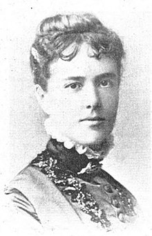 A white woman with her dark hair in an updo with a short curly fringe; she is wearing a high-collared dress with ruffles.