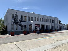 A gray art deco-style building with a black sparrow imprint on it. Yucca plants in planters stand in front of the building.