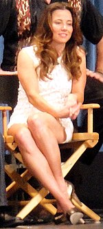 Linda seated with group, in director's chair, in white sleeveless mini and silver closed heels
