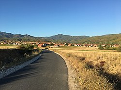 View from a single-track tarmac road running through fields or meadows of a village with red-roofed buildings with forested mountains in the background