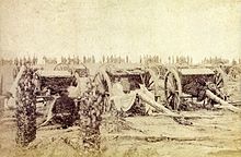 An old photograph showing a group of field artillery pieces and caissons with a line of soldiers in the background