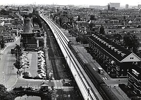 Photographie en noir et blanc et vu du dessus d'un paysage urbain traversé par une voie de chemin de fer longée à gauche par des bâtiments, un parking et un moulin à vent.