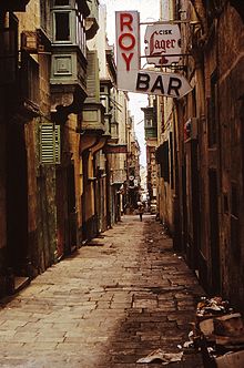 A narrow street with signs advertising "Roy Bar" and Cisk Lager.