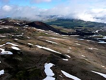 A sparsely snow-covered rocky landscape with a dark volcanic cone in the foreground and a cloud-covered mountain ridge in the background.