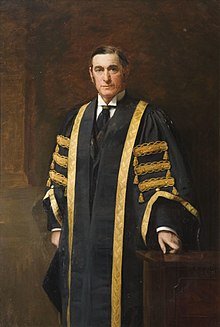 Three quarter length standing portrait, the subject looking directly at the viewer, while turned slightly to left. One hand rests on a low wooden table and he wears gowns which are black with a gold trim and details. Standing against a brown background.