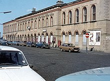 A white neo-Victorian station building, with a street full of parked 1970s cars in front of it.
