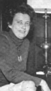 A candid photo of a smiling middle-aged white woman, seated, wearing a dark turtleneck sweater; her elbow is resting on her knee, and a lamp is visible by her side