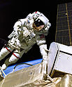 An astronaut in a white spacesuit with a red, white, and blue flag on the left shoulder in the foreground with a black background