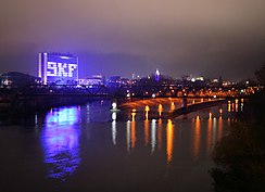 Allgemeine Dunkelheit. Im Vordergrund Wasser. Dahinter eine rechts orange beleuchtete Eisenbahnbrücke, im Hintergrund eine Skyline mit einem links herausstechenden blau beleuchteten rechteckigen Haus mit der Beschriftung „SKF“.