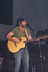A bearded man wearing a black baseball cap back to front, a green t-shirt and blue jeans, playing a guitar and singing into a microphone