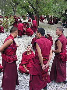 Groupe d'hommes, assez jeunes, vêtus d'une longue robe rouge. Certains sont debout, d'autres assis par terre, sur un sol caillouteux.