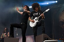 Singer and band performing on a dark stage in spotlight They are dressed in black; one plays a white guitar.