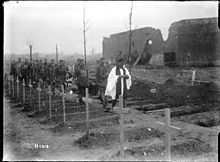 A priest leading soldiers carrying the coffin in a Belgian cemetery. There is a row of wooden crosses in the foreground with a collection of soldiers following the priest in single file.
