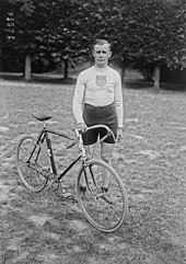 Photographie en noir et blanc d'un cycliste en short tenant un vélo par son guidon.