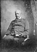 An older, balding black man with mustache, wearing a double-breasted suit and striped bow tie. He is sitting in a chair holding an open book in his left hand.