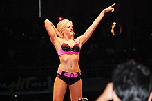 A blonde woman poses on the turnbuckles, while pointing forwards with one hand. She is wearing a black and pink crop top, and black shorts with a pink belt.