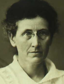 An older white woman, wearing pince-nez eyeglasses. Her hair is short and starting to grey at the temples. She is wearing a white blouse or dress with a wide collar.