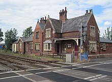 View across the tracks and level crossing of a fine 19th century station building in red brick and with stone mullioned windows, under a roof of Welsh slate. The platform has gone.