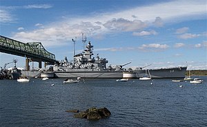 A large ship resting on the water under a bridge, smaller boats are visible in the foreground along with a ripple effect on the water.