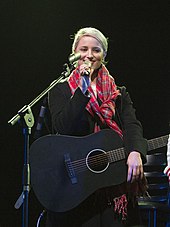 Дайанна Агрон, улыбаясь в теплом пальто и шарфе, держит микрофон и черную гитару на сцене.