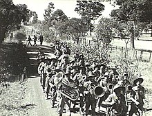 Soldiers marching along a dirt road in the Australian bush, led by a brass band