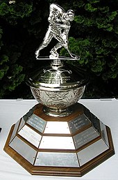 Silver-plated trophy including a figure of a hockey player mounted on a silver bowl above a hexagonal wooden base with nameplates