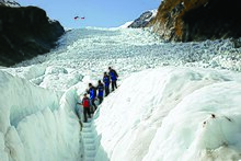 photograph of five people walking in a row on a snowy glacier with an helicopter visible in flight in the background