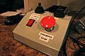 A closeup of a handmade electronic control box on a control room desk. The box has a slanted front–top face equipped with a red rocker switch labeled "3 degree Override" and a large red button labeled "Emergency STOP". The horizontal top face of the box has two identical small black joysticks with rubber bellows, labeled with directions. Some of the labels are hidden and/or blurry, but the left joystick appears to be labeled "UP/DOWN/LEFT/RIGHT" and the right joystick appears to be labeled "S/N/E/W" (compass directions rotated 180° from the usual arrangement). The box's sides are blue and its face is gray. The labels were printed by a label maker, and have black text on a white background.