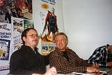 Photographie de deux auteurs de bande dessinée assis à une table pour dédicacer un livre.