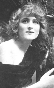 A young white woman with light hair, wearing a dark, one-shouldered dress