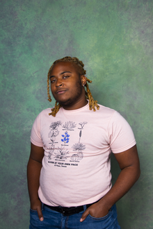 KB Brookins, a Black American writer with brown skin, poses with their hands in their pockets. They are wearing a pink shirt, blue jeans, and a Black belt. The background is purple and green.