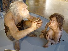 An old man is sitting to the left holding a stone blade, and a child is watching on the right, both clothed with only a wrap around the waist