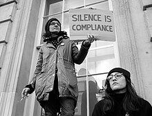 A protestor with a placard reading "Silence is Compliance" Silence is compliance - A protester with a message standing on a window ledge in Whitehall. (31903348794).jpg