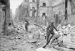 Two soldiers in a rubble-filled street bordered by badly-damaged buildings; one is clambering over the debris with a young child on his shoulder