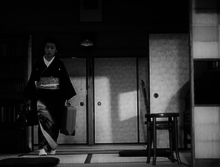 A middle-aged Japanese woman, wearing a kimono and carrying a suitcase in her left hand and a valise in her right, in the process of walking around the perimeter of a small room. There is a bookcase on the left and a straight-backed chair on the right and screens and a ceiling lamp in the near background.