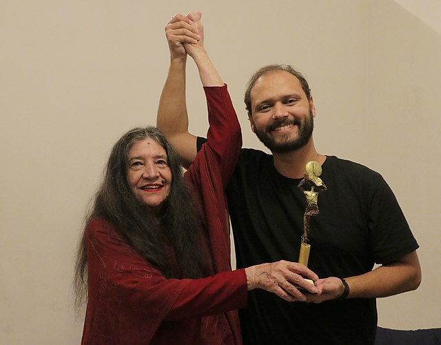 Da esquerda para a direita: Nisete Sampaio de vermelho sorrindo com o troféu do festival na mão junto do cineasta Thiago Prado.