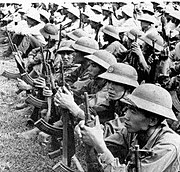 Большое количество хорошо экипированных азиатских солдат сидят рядами на земле, в пробковых шлемах и с автоматами в руках.