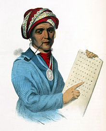 一个北美原住民男人，头上戴着镶银边的红色头巾，嘴叼烟斗，胸前挂着银奖牌，身穿青蓝色上衣，左手拿着写有切罗基音节文字的平板，右手指着平板上的文字。