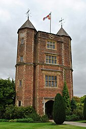 a high tower of red brick with two pyramid roofs and a flag flying