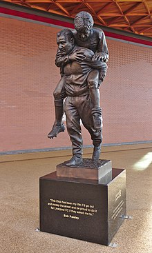 Photo d'une statue présentant un homme âgé portant un joueur de football, visiblement blessé, sur son dos.