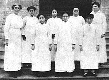 Eight women, standing, all wearing nurses' white uniforms. The middle six women are young Chinese nursing students; the white woman on the left of them is tall with greying hair; the white woman on the right of them has dark hair and a plump body.