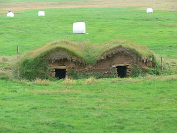 Paysage agricole : anciens bâtiments en terre à toiture végétale, et balles d'herbe enrubannée dans leur emballage blanc