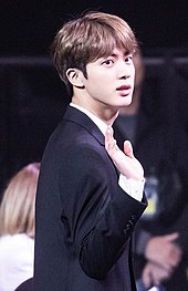 Jin, wearing a formal black suit at an award ceremony, waves to the crowd.
