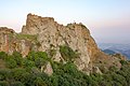 Sur un rocher aux teintes grises et rouille, parsemé de quelques buissons, s'élèvent des ruines desquelles émerge une croix.