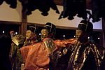 Three masked men in traditional Japanese clothes, one in orange the others in green gold.