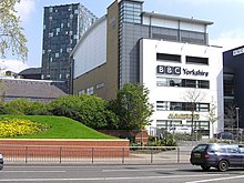 A quirky modern five-storey building with a large sign saying "BBC Yorkshire" in black above the second-floor windows on the white-fronted façade of the lower four floors can be seen on the far side of a dual-carriageway road with a barrier along the central reservation. At right-angles to the right of the building is a tall blue slab with the letters "BBC" in white at the top. The left side of the building is mostly brick-red with a few windows, but above it is a light blue windowless section. The roof above this and the grey fifth floor of the frontage curves gently down to the rear. A lone car is driving from left to right along the road; between it and the building, temporary boards have been erected in front of a building to the left. In the top left-hand corner of the picture, part of a tall many-windowed building can be seen.