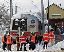 Eight men wearing neon orange jackets or vests with reflective stripes and green or white hard hats, most with their backs to the camera, stand in a ragged row in snow across the bottom of the image. In front of them is the front of a gray rail passenger train with a wide red stripe around its lower section between two gray square metal columns that rise to the top of the image. The words "Metro-North Commuter Railroad" and the Connecticut state seal are visible on one side of the front. Its roof is covered with snow. In the rear right is a dull yellow off-center snow-covered gabled canopy with the words "New Canaan" in the field sheltering a concrete platform.