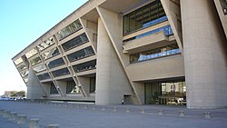 A tall beige building with an angled front face, leaning out from the top, is supported by three columns and covered with rows of windows