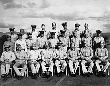 Двадцать восемь моряков в форме ВМС США позируют на палубе авианосца времен Второй мировой войны.