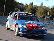 Photo de la Citroën Xsara WRC de Sébastien Loeb, livrée bleue et rouge, vue de face sur une route goudronnée.